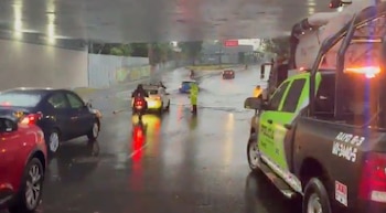 Vista de un paso a desnivel inundado por la lluvia intensa, con varios coches parados o avanzando lentamente, una patrulla policial y personas en el agua