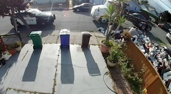 Vista aérea de un camión volcando basura en un jardín residencial junto a una valla de madera, con un coche de policía y tres cubos de basura vacíos en primer plano