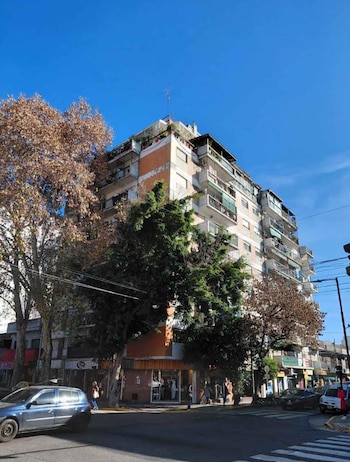 Edificio de apartamentos de múltiples pisos, árboles con follaje verde y seco, vehículos estacionados y en circulación, personas caminando en una esquina urbana