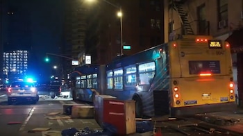 Imagen nocturna de un autobús articulado de la MTA en una calle urbana. Hay un coche de policía con luces azules y escombros de un comedor exterior