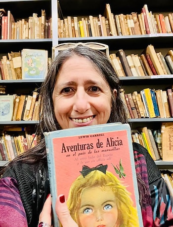 Mujer de cabello gris y gafas en la cabeza sonríe, sosteniendo un libro de "Aventuras de Alicia" con portada de niña rubia, frente a estanterías llenas de libros