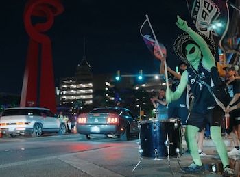 Un aficionado vestido de alienígena verde con un sombrero y camiseta de los Spurs toca un tambor en la calle por la noche, con coches y edificios de fondo