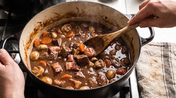 Una persona mezcla un estofado en una olla de hierro fundido con carne, panceta, champiñones y zanahorias usando una cuchara de madera. Se observa vapor.