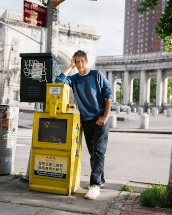 Un hombre con sudadera azul y jeans oscuros se apoya en un dispensador de periódicos amarillo. En el fondo, un arco y un edificio, con un cartel de tráfico