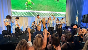 Banda en un escenario con camisetas de Argentina frente a una pantalla con un partido de fútbol. El público, en primer plano, levanta las manos celebrando