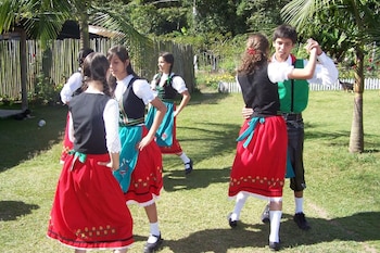 Jóvenes vestidos con trajes folclóricos austro-alemanes, con faldas rojas y chalecos oscuros, bailan sobre césped verde en un día soleado