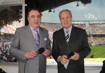 Dos hombres con trajes y corbatas sostienen micrófonos dentro de una cabina, con un estadio de fútbol lleno de público y el campo verde visible al fondo
