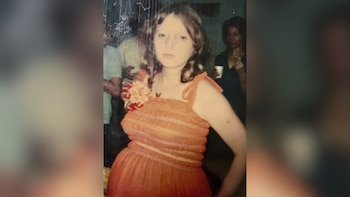 Una mujer joven con cabello castaño claro y un vestido de tirantes naranja con adornos de flores en el hombro izquierdo, posa frente a un fondo borroso con otras personas