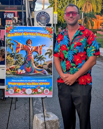 Un hombre con camisa floreada y lentes oscuros está de pie junto a un cartel con la ilustración de una estatua colorida de Celia Cruz