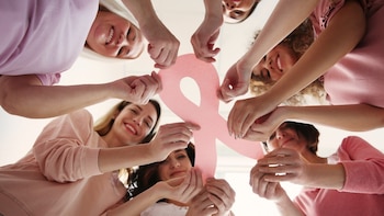 Varias mujeres dispuestas en círculo sostienen juntas un gran lazo de papel rosa visto desde un ángulo inferior