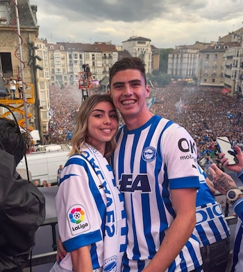 Hombre y mujer sonríen en primer plano con camisetas de fútbol a rayas azules y blancas, frente a una multitud en una plaza urbana con edificios