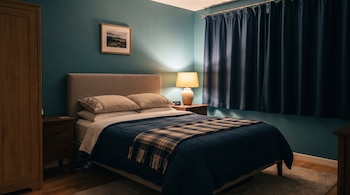 Una cama vestida con azul, dos mesas de noche, una lámpara encendida, cortinas oscuras cerradas y un armario de madera.