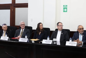 Cinco personas, tres hombres y dos mujeres, sentadas en una mesa larga con placas de identificación, micrófonos, botellas de agua y señal de evacuación
