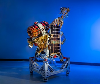 Modelo de nave espacial o satélite con aislamiento dorado y paneles solares negros, montado sobre una estructura de metal gris. Fondo azul liso