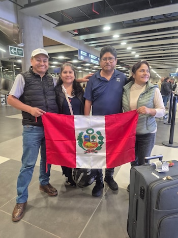 Cuatro personas, dos hombres y dos mujeres, posan en un aeropuerto. Sujetan una bandera de Perú. Hay una maleta gris y señalización de aeropuerto