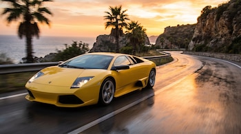 Un Lamborghini Murciélago amarillo circula por una carretera costera mojada y curva. Palmetas y acantilados rocosos bordean la carretera con el océano y un cielo naranja de atardecer de fondo.