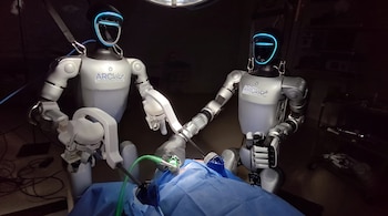 Dos robots grises con el logo ARCLab y luces azules en la cabeza operan sobre una sábana quirúrgica azul con instrumentos médicos y cables verdes.