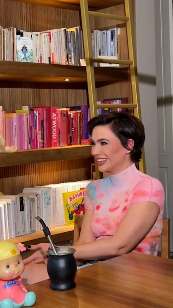 Una mujer con cabello corto y blusa de colores sonríe sentada frente a una mesa con un mate y una biblioteca al fondo