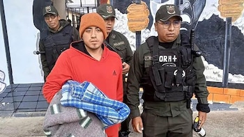 Un hombre con gorro naranja y sudadera roja es escoltado por dos policías uniformados con chalecos tácticos. El hombre sostiene una manta doblada