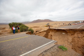 Carretera asfaltada con tramo colapsado por un barranco de tierra. Personas observan, una excavadora amarilla, camiones y automóviles. Montañas y cielo nublado