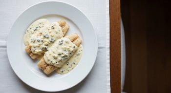 Vista cenital de un plato blanco con dos filetes de merluza, cubiertos con salsa cremosa de Roquefort, sobre un mantel blanco en una mesa de madera oscura.