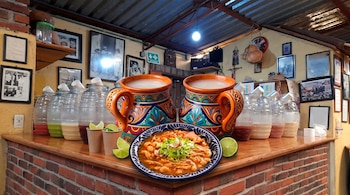 Un plato de pozole, dos jarras de barro con motivos coloridos, limones y recipientes de vidrio con bebidas sobre un mostrador de ladrillo.