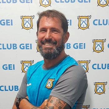 Hombre con barba y tatuajes, camiseta azul y gris, brazos cruzados, frente a una pared blanca con logos y texto Club GEI