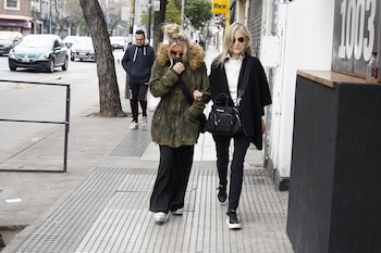 Dos mujeres caminan por una acera de ciudad. Una con chaqueta verde y gafas de sol, otra con poncho negro y gafas oscuras, junto a árboles y edificios