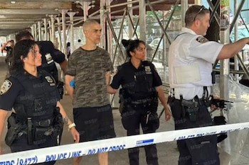 Tres agentes de policía, dos mujeres y un hombre, escoltan a un hombre con esposas azules junto a una cinta policial. Se ve un andamio de fondo