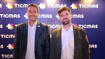 Dos hombres sonrientes en blazers posan frente a un muro azul con el logo repetido de Ticmas. El de la izquierda lleva blazer oscuro, el de la derecha blazer marrón