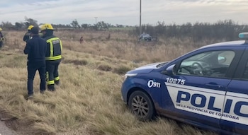 Una patrulla de policía de Córdoba azul y blanca y dos rescatistas en uniforme amarillo en la hierba seca, con un vehículo blanco siniestrado al fondo