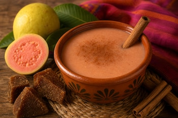 Tazón de barro con atole y una rama de canela, junto a guayabas frescas, trozos de piloncillo y ramas de canela sobre una mesa.