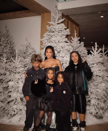 Kim Kardashian sonríe con cuatro niños de pie frente a ella, todos vestidos de oscuro, rodeados de varios árboles de Navidad blancos con luces