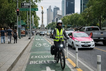 Persona con casco y chaleco reflectante en bicicleta eléctrica por ciclovía verde en CDMX. Se ve el Ángel de la Independencia, edificios y tráfico urbano al fondo.