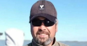 Primer plano de hombre de mediana edad sonriendo, con gorra oscura, gafas de sol y barba canosa, con un cielo azul y agua de fondo