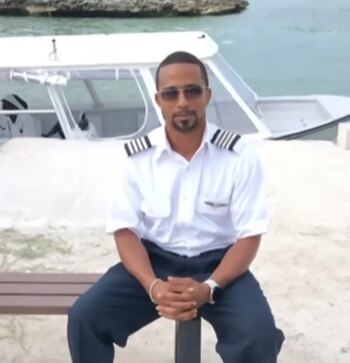 Hombre con barba, gafas de sol y uniforme de piloto (camisa blanca, charreteras), sentado en un banco. Detrás, una lancha blanca y el mar