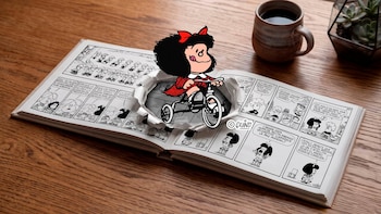 Cómic abierto sobre mesa de madera. Mafalda en color sobre triciclo emerge de una hoja rasgada del cómic. Taza de café y planta suculenta
