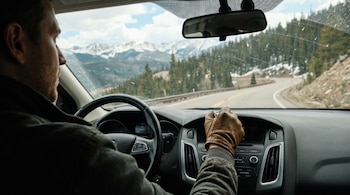 Vista interior de un coche. Se ve el perfil de un conductor y su mano enguantada sobre el salpicadero. Al frente, una carretera sinuosa y montañas nevadas.