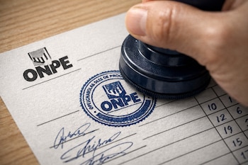 Una mano sostiene un sello azul oscuro estampando un logo de la ONPE sobre un acta electoral blanca con texto, firmas y números, sobre una mesa de madera clara.