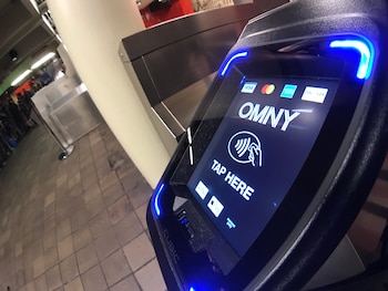 Terminal de pago OMNY con pantalla azul. Muestra logo OMNY, icono de pago sin contacto, 'TAP HERE' y logos de tarjetas de crédito y monederos digitales