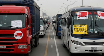 Larga fila de camiones de carga rojos, azules y buses blancos y azules bloquean una avenida principal bajo un cielo nublado. Varios vehículos exhiben carteles de protesta.