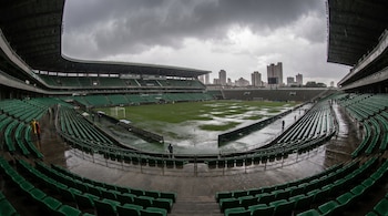 Estadio de fútbol con gradas verdes, campo de juego inundado por la lluvia, personas con impermeables y edificios al fondo bajo un cielo nublado.