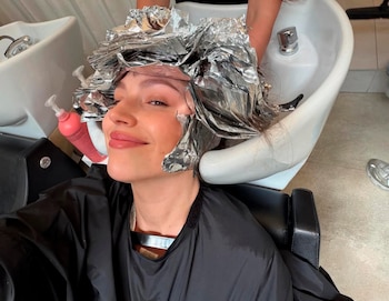 Úrsula Corberó en un salón, sonriendo, con el cabello envuelto en papel de aluminio para un tratamiento de coloración y una capa protectora negra