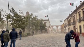 Vista de una plaza en Querétaro con personas abrigadas, árboles meciéndose y una tolvanera densa. Banderas mexicanas ondean en edificios y un acueducto es visible al fondo.