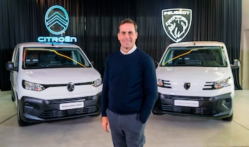 Un hombre sonriente en el centro, vestido con un suéter azul y pantalón gris, flanqueado por dos furgonetas blancas idénticas. Detrás, logos de Citroën y Peugeot