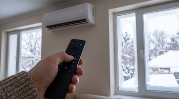 Una mano sostiene un control remoto y apunta a un aire acondicionado en una pared. Al exterior, a través de dos ventanas, se ve un paisaje nevado de invierno.