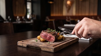 Un corte de carne roja sobre una tabla de madera junto a patatas asadas y una mano que sostiene un tenedor y un cuchillo.