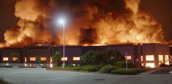 Vista nocturna de un gran almacén ardiendo intensamente, con llamas anaranjadas y humo denso sobre el edificio, y una farola iluminando el primer plano