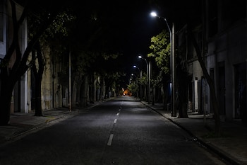 Calle de asfalto vacía durante la noche con veredas, árboles y farolas encendidas a los lados.