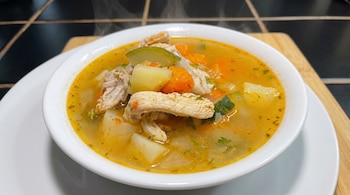 Un tazón blanco lleno de caldo de pollo caliente con vapor. Contiene trozos de pollo deshebrado, patatas, zanahorias y calabacín. Servido sobre una tabla de madera.